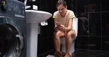 Young woman uses mobile phone sitting on toilet at home. Gadget addiction and social media browsing concept