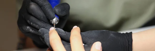 Close-up of nail master using electrical equipment to clean client nails in modern beauty studio. Worker wear protective black gloves. Self care concept