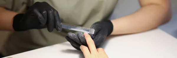 Close-up of nail master using nail file tool to create perfect shape for clients nails. Worker wear protective black gloves for work. Beauty studio concept