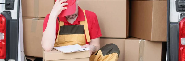 Close-up of delivery man sit in van with delivery receipt and wait for client. Middle aged courier in red uniform. Fast shipping, address delivery concept