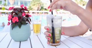 Havuzun arka planında yaz kokteyli olan kadın elleri, yakın plan. Kumsaldaki kafede, masada güzel bir çiçek.