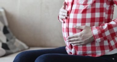 A pregnant woman in a red plaid shirt strokes her belly, close-up. Late pregnancy, communication with the baby before birth