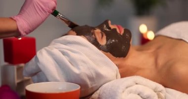 Beautiful woman lies with eyes closed and enjoys rejuvenating and moisturizing spa treatment in beauty salon. Hand of professional cosmetologist applies black clay mask to client face