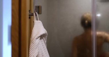 Naked woman washes in shower white bathrobe hangs on hook. Body hygiene and showering