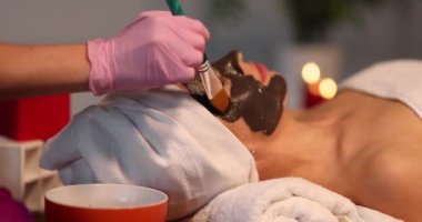 Cosmetologist brushing a chocolate mask on the face of a lying woman, slow motion. Relaxing salon treatment
