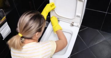 A woman in yellow gloves wipes a white toilet bowl at home, close-up, back view. Black bathroom cleaning