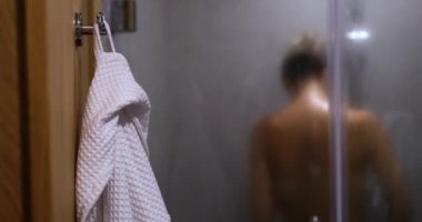In the foreground, a bathrobe hangs in the bathroom. Woman washing herself in the shower, view from the back