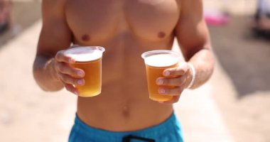 Hands hold two plastic glasses with beer on the beach in summer. Concept of cultural consumption of beer among tourists