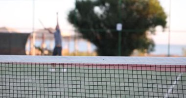 Through net portrait of man playing tennis on court outdoors. Player tennis player hits ball on tennis court concept