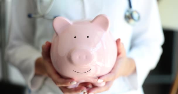 医生女士牵着并捂住笑容满面的小猪银行 手牵手进行特写 保健保险的经济学 — 图库视频影像