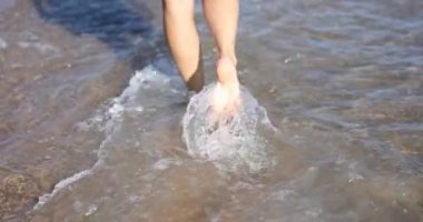 İnce kadın bacakları ve ayakları kumlu sahilde deniz suyu dalgaları üzerinde yürüyor. Deniz sörfünde yürüyen güzel bir kadın