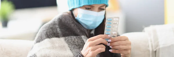 Sick woman in a mask sits in a blanket on the couch, high temperature, close-up. Medicines to treat the virus