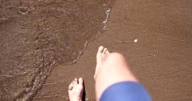 Deniz kumu üzerinde ayaklar ve dalgalar kıyı şeridi boyunca ilerler. Okyanus sahili tatili ve yaz turizmi