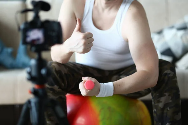 Athlete man holds thumbs up and raises dumbbells sitting on ball at home in front of camera. Recommendations for home fitness and sports training remotely