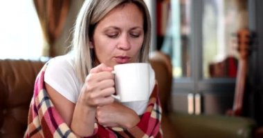 Grip olmuş genç bir kadın öksürdüğü için bir fincan sıcak çaya üflüyor. Soğuk kış konsepti ve rahat ev