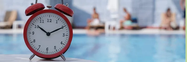 Red alarm clock standing on edge of hotel swimming pool closeup. What time can you sunbathe concept