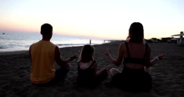 Anne baba ve kızı gün batımında plajda yoga yapıyorlar. Mutlu dost aile ve rahatlama