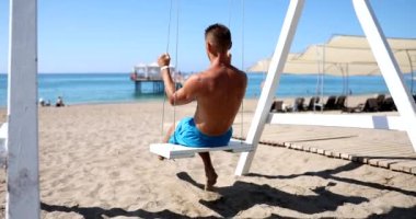 Genç gezgin adam deniz kenarında salıncakta yaz tatilinin tadını çıkarıyor. Plajda sallanarak turistleri rahatlatmak ve seyahat etmek.