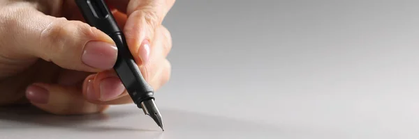 Female fingers hold a black fountain pen on a gray background, close-up. Correspondence concept, copywriting