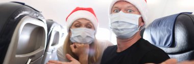 Koruyucu tıbbi maskeli üzgün ve sarhoş adam ve kadın ve Noel Baba kırmızı şapkalı uçaklar. Covid19 salgını sırasında yeni yıl yolculuğunun zorlukları