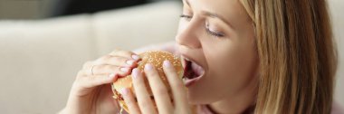 Genç sarışın kadın portresi sulu hamburger yiyor, abur cubur yiyor, yağlı öğle yemeği yiyor, eve fast food siparişi veriyor. Sağlıksız yemek, paket servis, açlık.