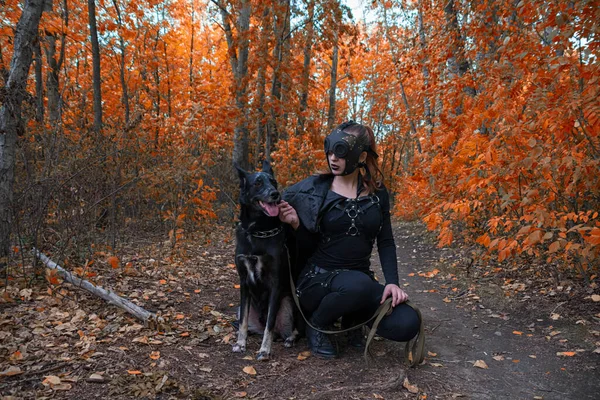 Uma menina em um traje bdsm e uma máscara preta e com um cão na floresta vermelha.an uma ideia para o Halloween. — Fotografia de Stock