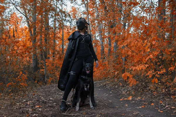 Uma menina em um traje bdsm e uma máscara preta e com um cão na floresta vermelha.an uma ideia para o Halloween. — Fotografia de Stock