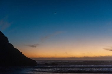 Pfeiffer sahilinde parlak turuncu gökyüzü, gün batımında. Arka planda küçük hilal var..