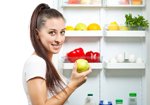 Menina bonito com a maçã perto do refrigerador aberto — Fotografia de Stock