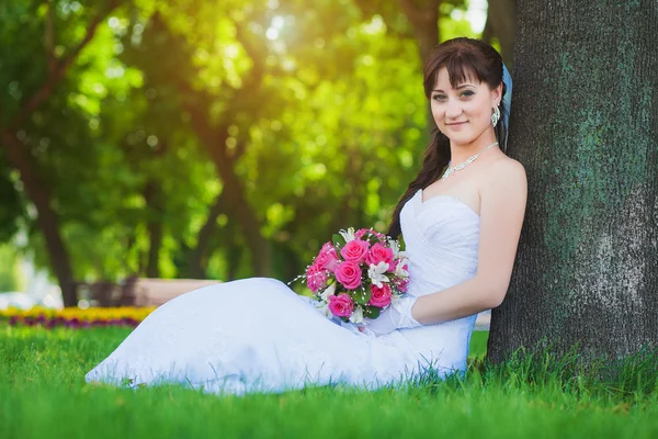 Beautiful bride in white dress sitting under a tree — Stock Photo, Image