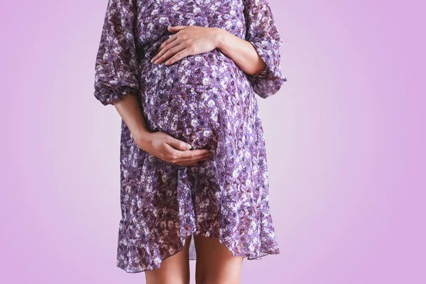 Pregnant woman in dress holds hands on belly on a violet background. — Stockfoto