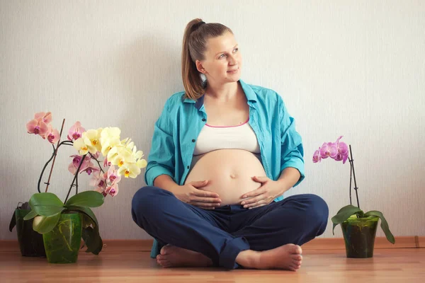 Gravid kvinna som sitter på golvet med orkidéblommor hemma. väntar på det nyfödda barnet. — Stockfoto