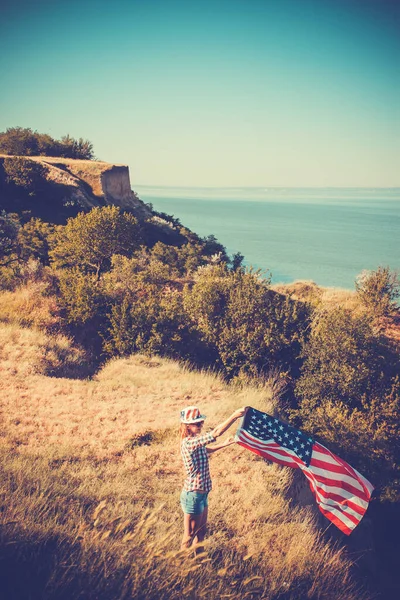 Hermosa joven sosteniendo una bandera americana en el viento en un prado. Paisaje de verano contra el cielo azul. —  Fotos de Stock