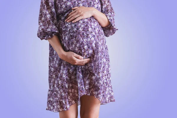 Těhotná žena v šatech drží ruce na břiše na fialovém pozadí. — Stock fotografie