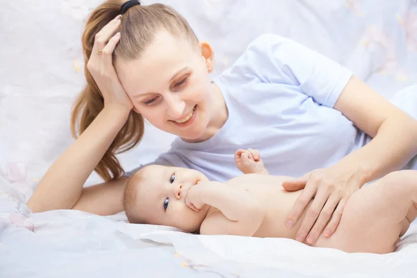 Young happy mother together with her newborn baby lie cheerfully on a white bed — Stock Photo, Image