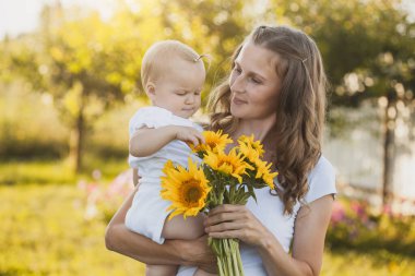cute one year old baby girl with mom in a blooming garden with a bouquet of sunflowers