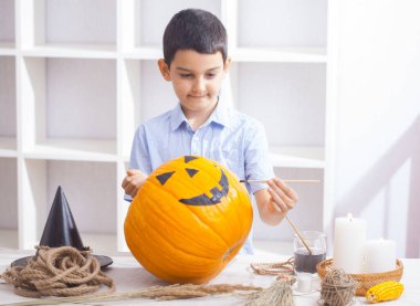 Boy in apron painting Halloween pumpkin, at wooden table, preparing holiday decorations at home