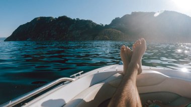 First person view of a man relaxing on a boat with his legs resting on the hull, against a seascape with islands