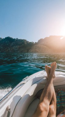 First person view of a man relaxing on a boat with his legs resting on the hull, against a seascape with islands