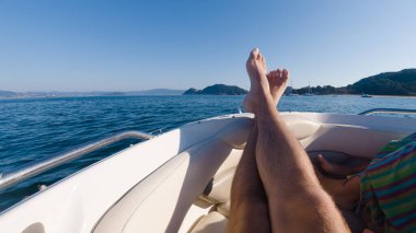 First person view of a man relaxing on a boat with his legs resting on the hull, against a seascape with islands