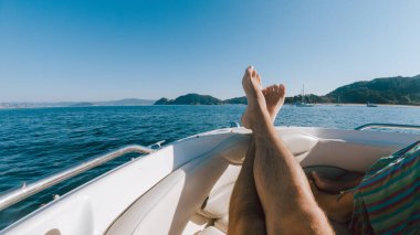 First person view of a man relaxing on a boat with his legs resting on the hull, against a seascape with islands