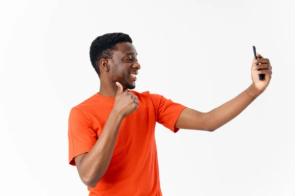 Afrika görünümlü ve turuncu tişörtlü bir adam başparmağını gösteriyor ve bir cep telefonuna bakıyor. — Stok fotoğraf