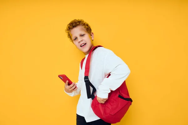 Fröhlicher Schuljunge in weißem Sweatshirt Handy roter Rucksack gelber Hintergrund — Stockfoto