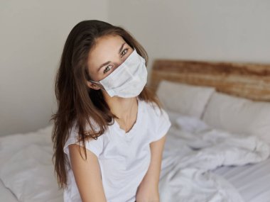 Tıbbi maskeli genç bir kadın başını yana eğdi ve yatağın içine oturdu.