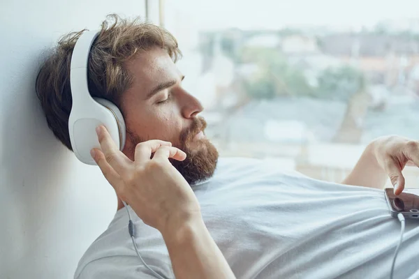 Χαριτωμένος άντρας ξαπλωμένος κοντά στο παράθυρο φορώντας ακουστικά διασκέδαση — Φωτογραφία Αρχείου