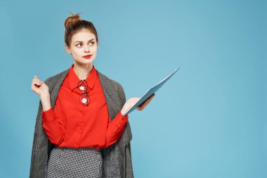 İş kadını, omuzlarında ceket, elinde klasörle mavi arka planda çalışıyor.