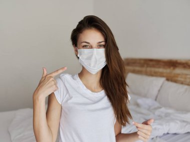 Tıbbi maskeli güzel bir kadın parmaklarını gösterip içeride yatakta oturuyor.