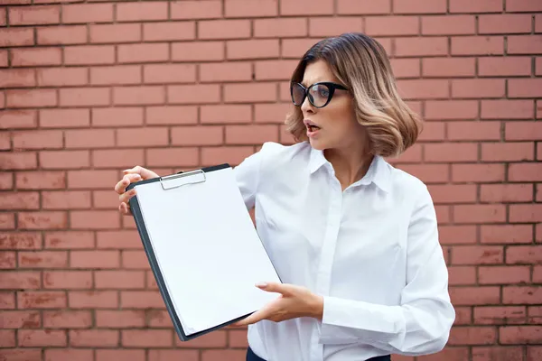 Femme d'affaires en chemise blanche et lunettes avec des documents sur le mur de briques de la rue — Photo