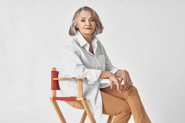 Starší žena v bílé košili sedí na židli a pózuje — Stock fotografie