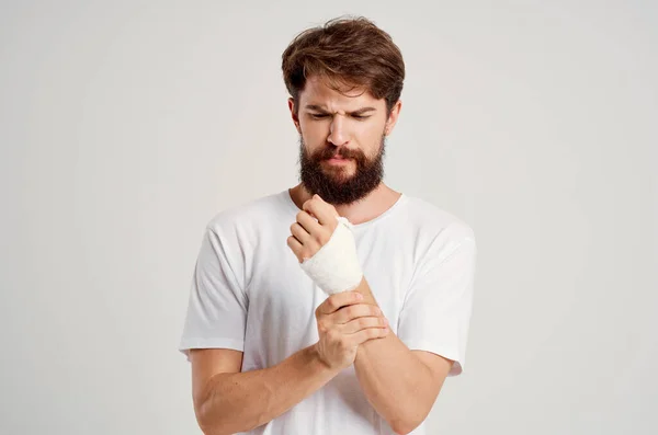 Hombre barbudo en una camiseta blanca con una mano vendada posando fondo claro —  Fotos de Stock